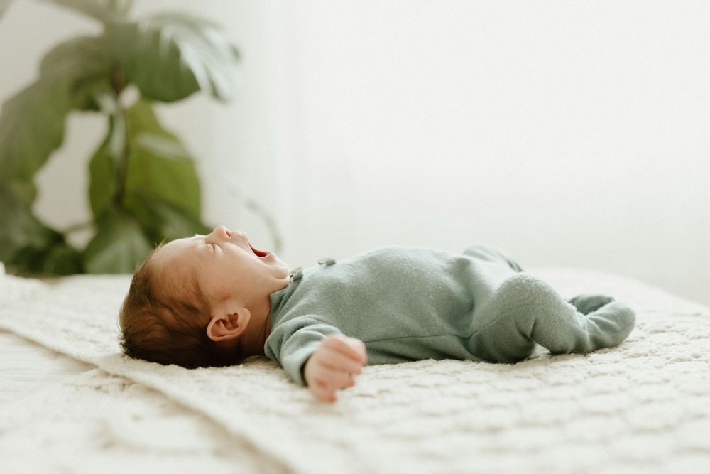 Newborn baby sleeps peacefully in a cozy gray swaddle on white bedding in dreamy natural light.