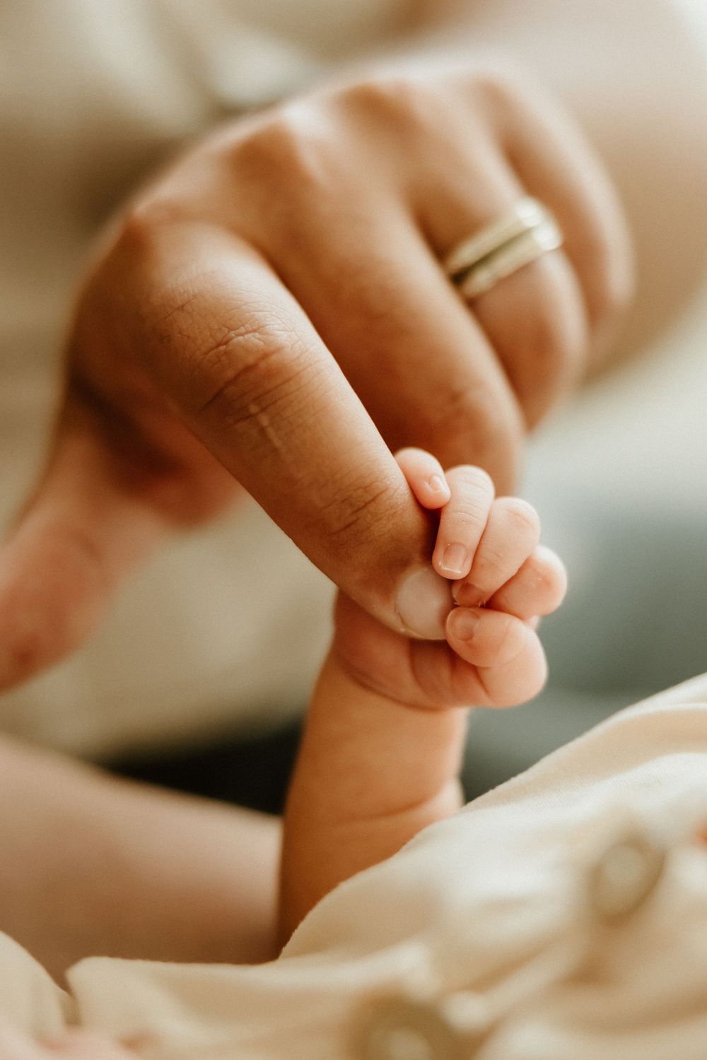 A tiny baby's hand grasps an adult finger wearing a wedding ring against a soft white background.