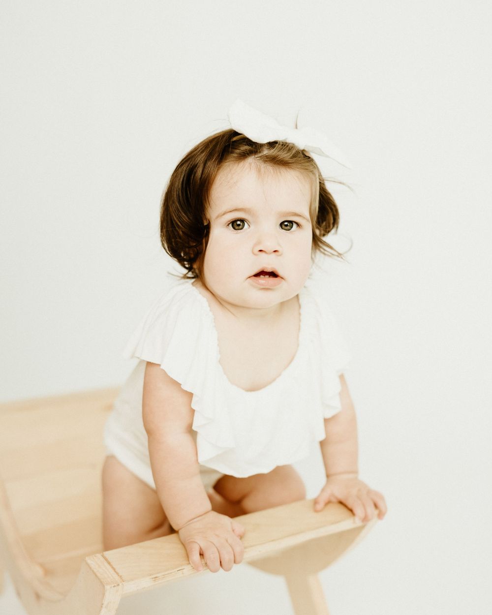 Small child in a white outfit sitting on a wooden surface against a light background.