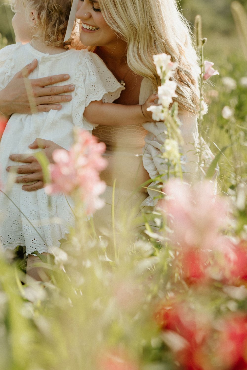 A tender embracing moment captured in a sunlit wildflower meadow with pink and coral blooms in the foreground.