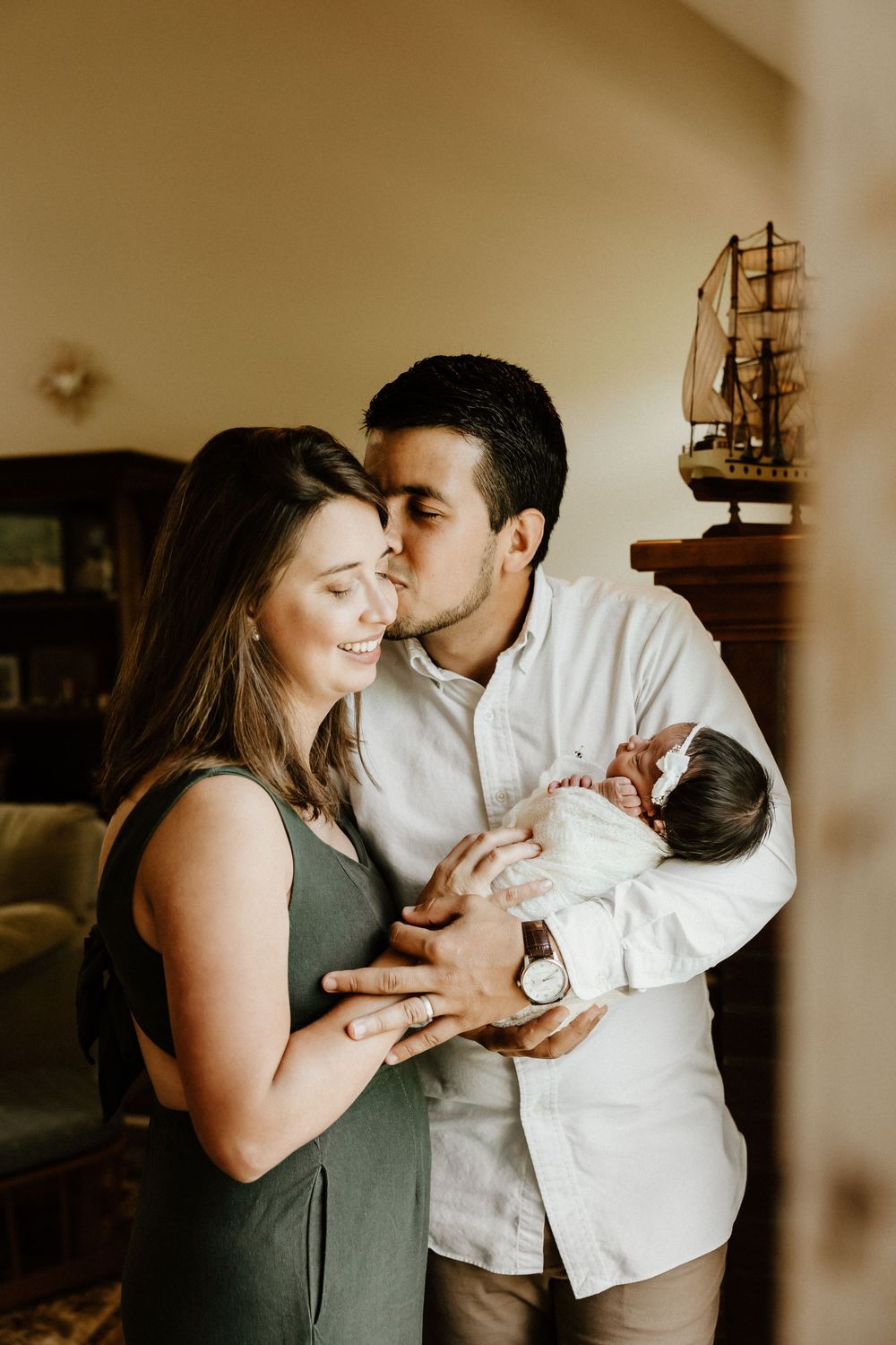 A tender family moment at home as parents hold their newborn baby, with a decorative model ship visible in the background.