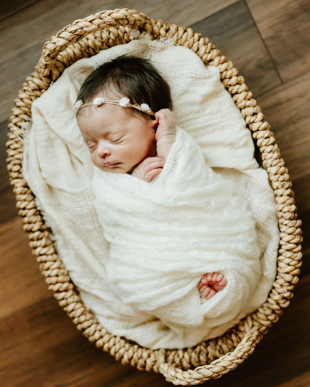 A sleeping newborn baby swaddled in white fabric lies peacefully in a woven basket on a wooden floor.