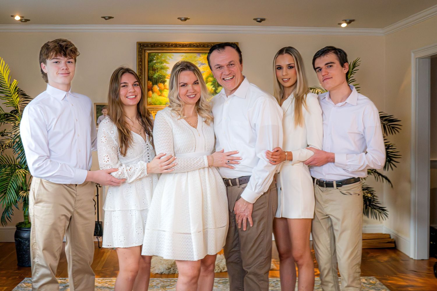 A group in coordinated white and beige outfits poses together in a bright indoor setting with tropical plants.