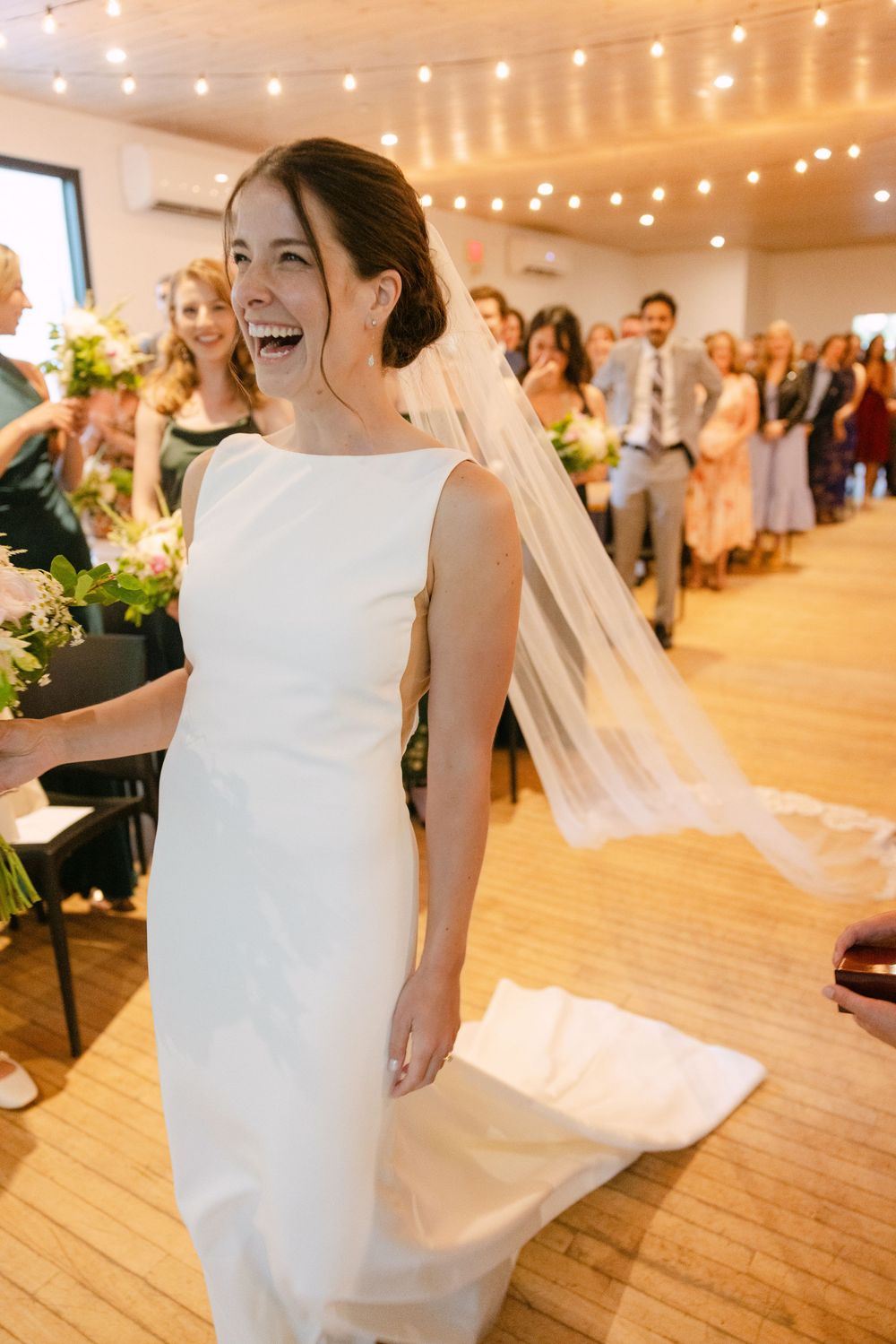 A joyful bride in a sleek white dress laughing during a wedding reception under string lights in a bright wooden room.