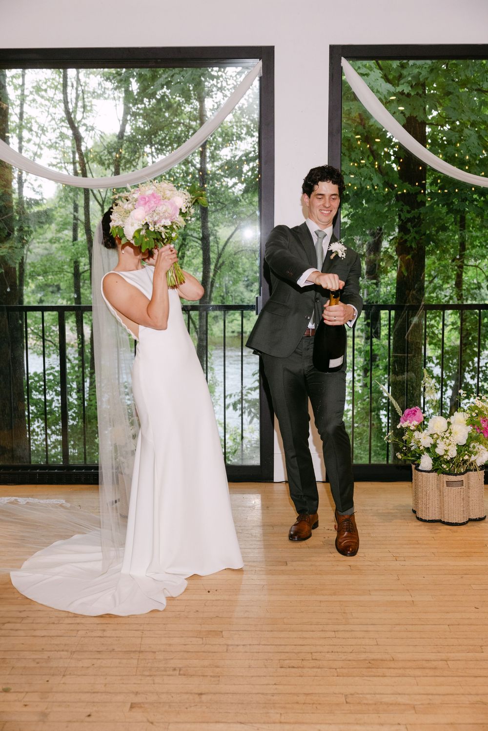 A couple in wedding attire celebrate with champagne on a wood-floored venue decorated with white draping and floral arrangements.