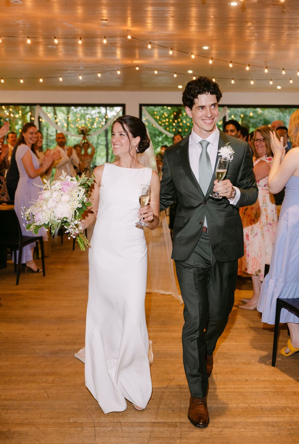 A happy couple walk down the aisle at their wedding reception under twinkling lights and greenery.