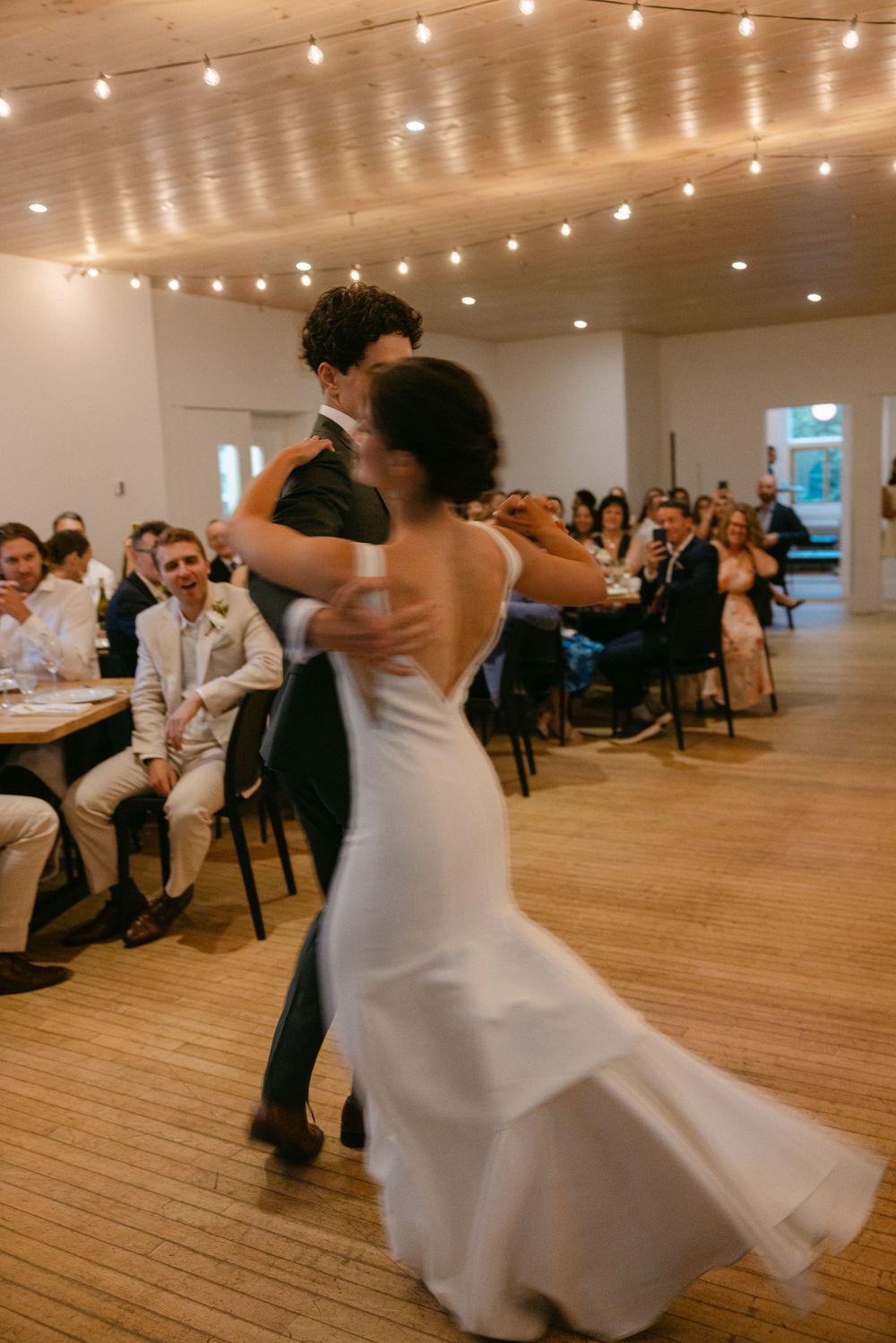 Wedding reception at indoor venue with string lights and guests dancing on wooden floor during romantic first dance.
