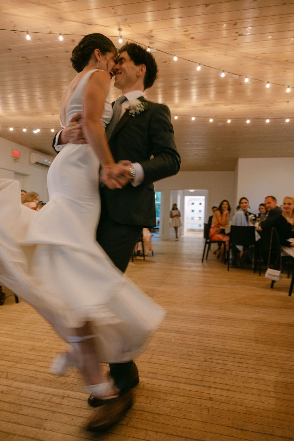 A couple dances elegantly in a rustic wooden hall with string lights hanging overhead, captured in a dynamic motion sequence.