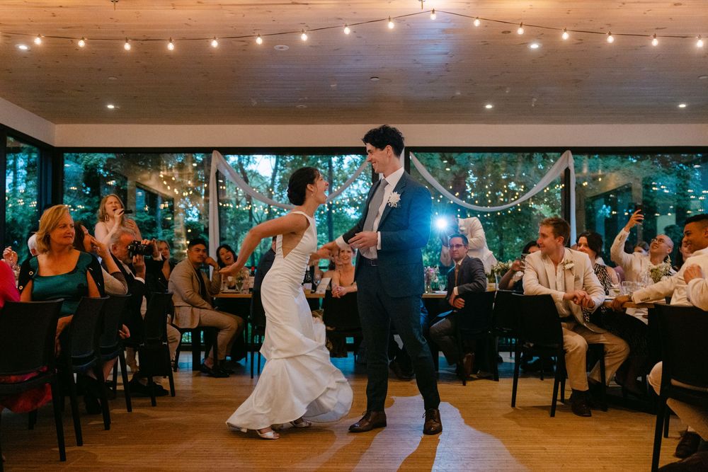 A couple shares their first dance under string lights at a wedding reception in a rustic venue while guests look on.
