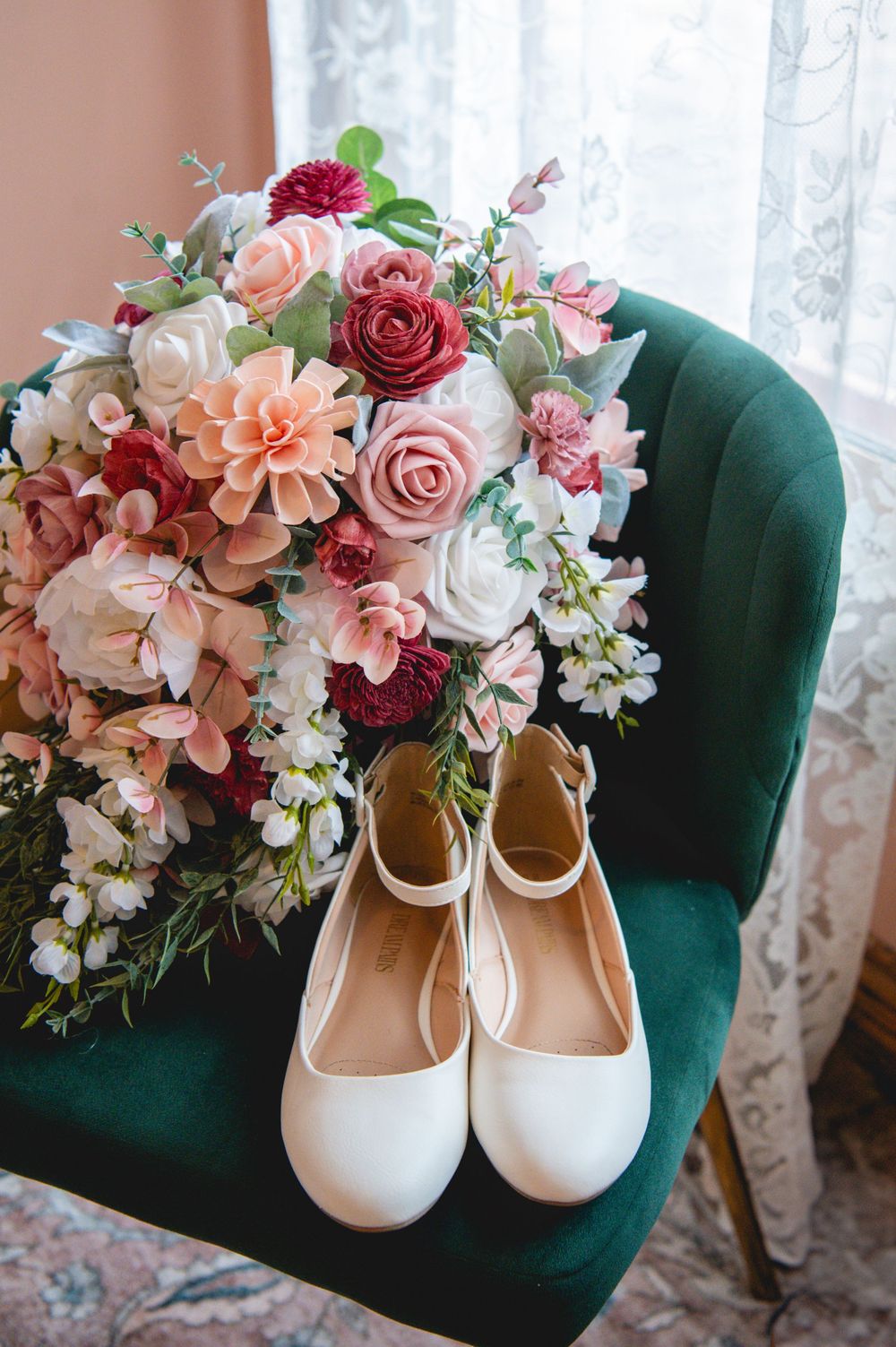 A romantic bridal bouquet of pink and burgundy roses sits beside white ballet flats on an emerald velvet chair.