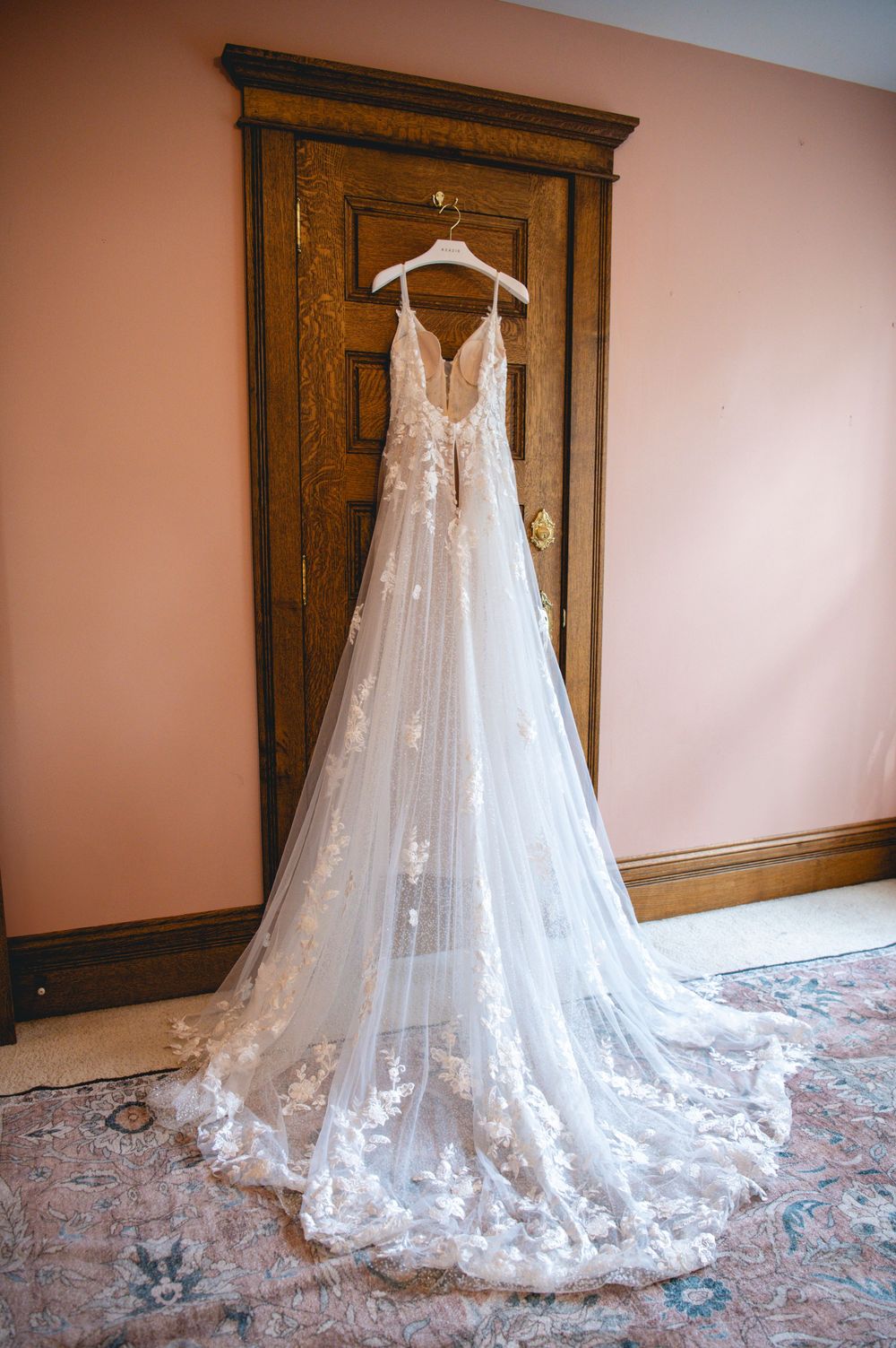 An elegant lace wedding dress with long train and deep V-neckline hanging on wooden door.