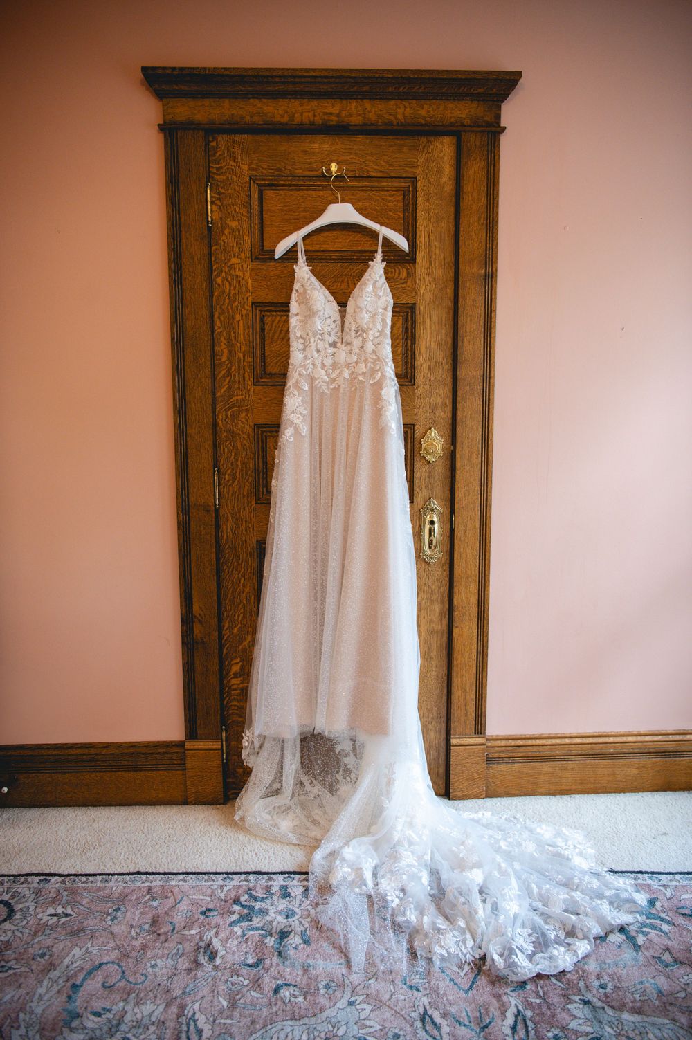 A delicate white lace wedding gown with long train hangs elegantly on a wooden door in soft lighting.