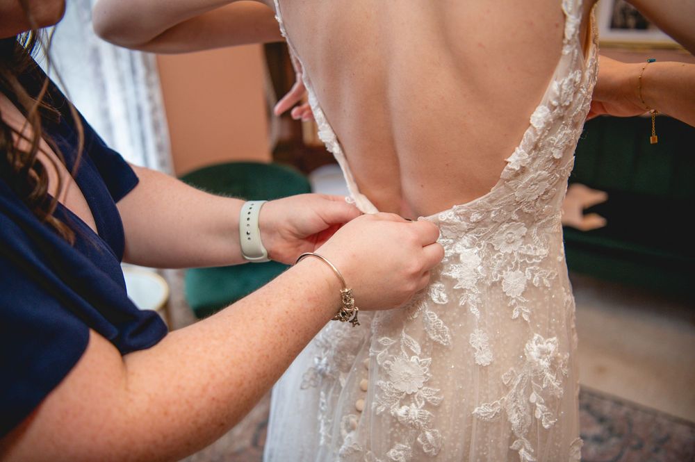 A delicate white wedding dress being buttoned up, showing intricate lace and floral embroidery details on the back.