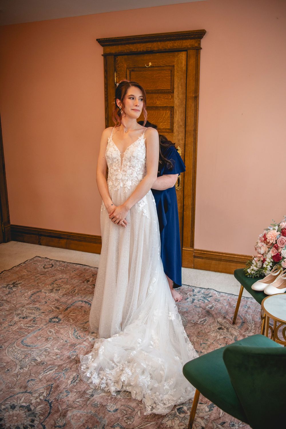 A bride wearing a flowing white lace wedding gown stands elegantly in front of wooden armoires on a patterned carpet.