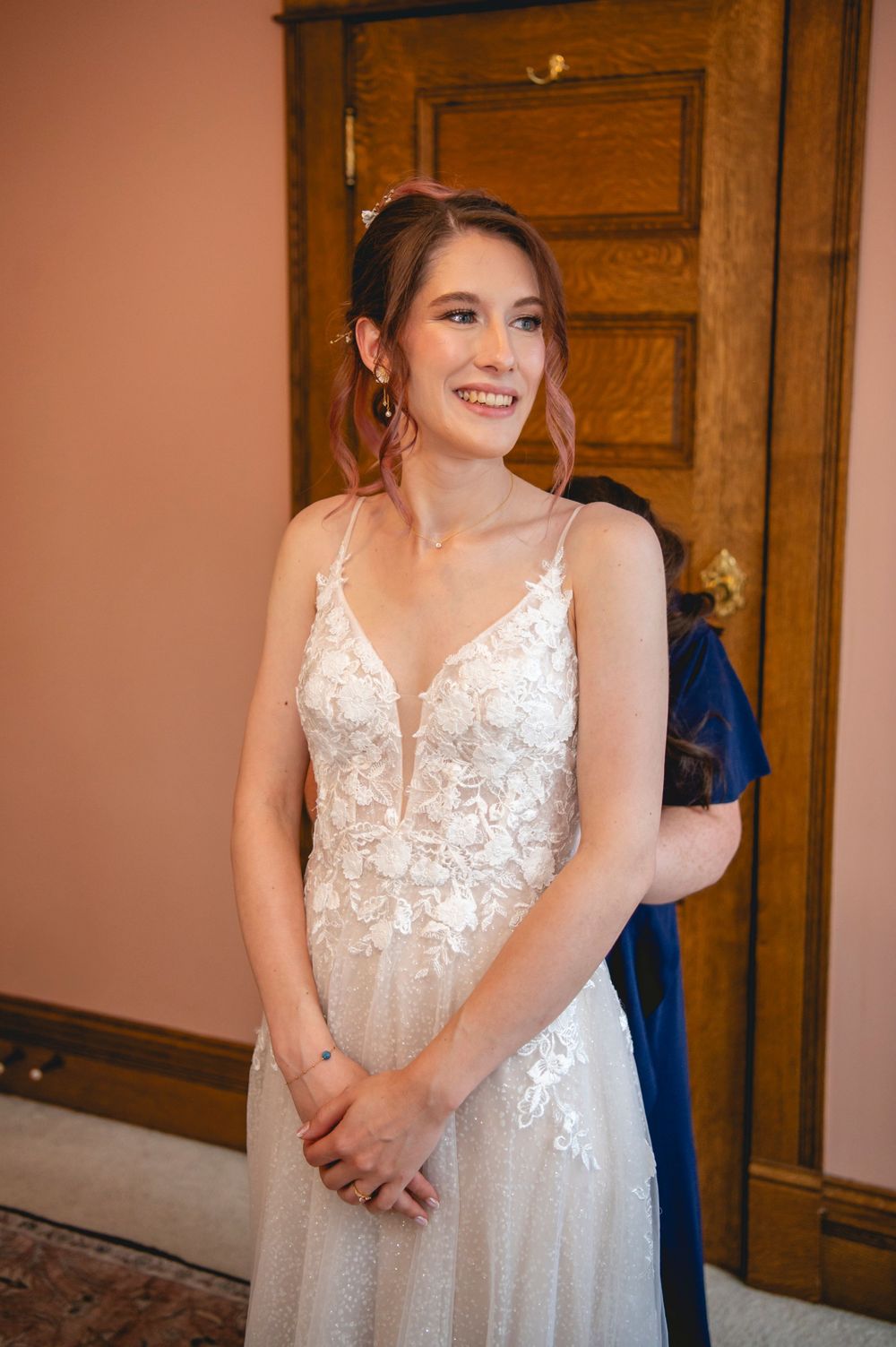 A woman wearing a delicate white lace wedding dress with spaghetti straps stands smiling in front of a wooden door.