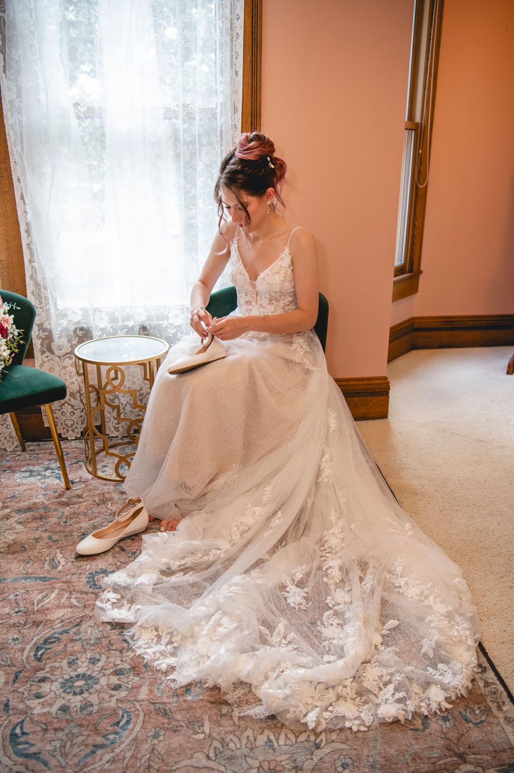 A person in a lace wedding dress with a flowing train sits elegantly in a room with sheer curtains and ornate decor.