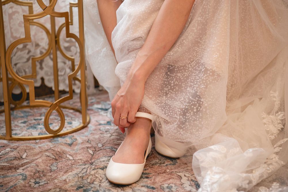 Close-up of a bride adjusting her white ballet flat shoe while wearing a sparkly wedding gown.