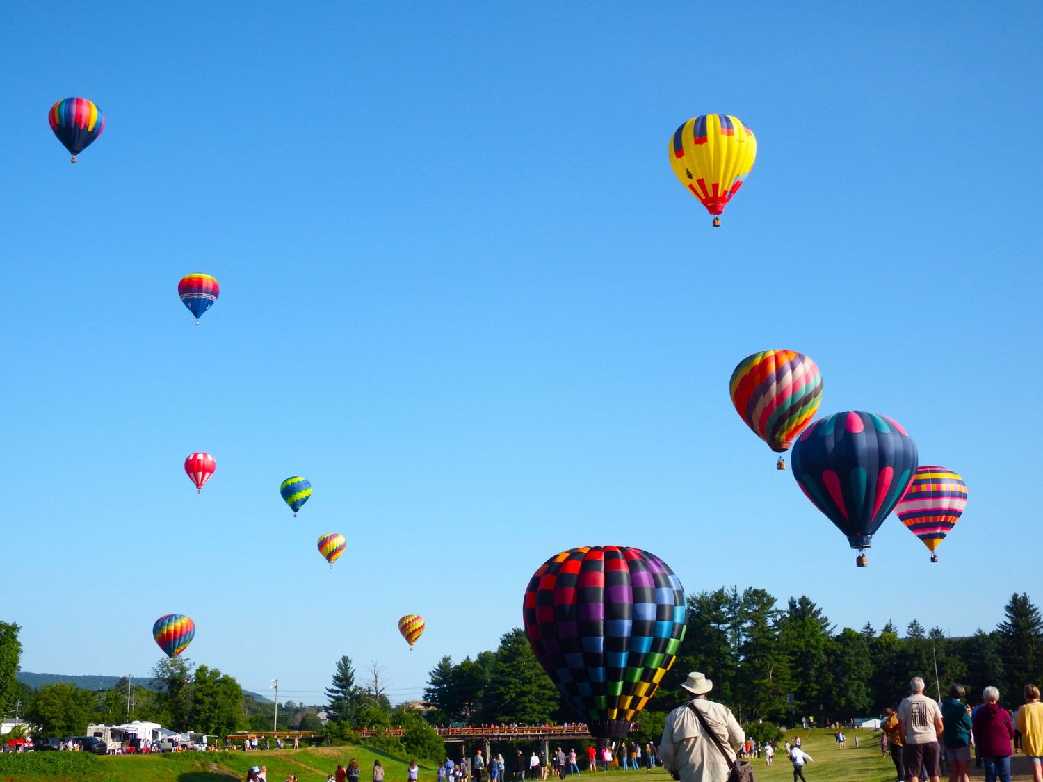 Colorful hot air balloons float in a bright blue sky during a festival against a scenic landscape backdrop.