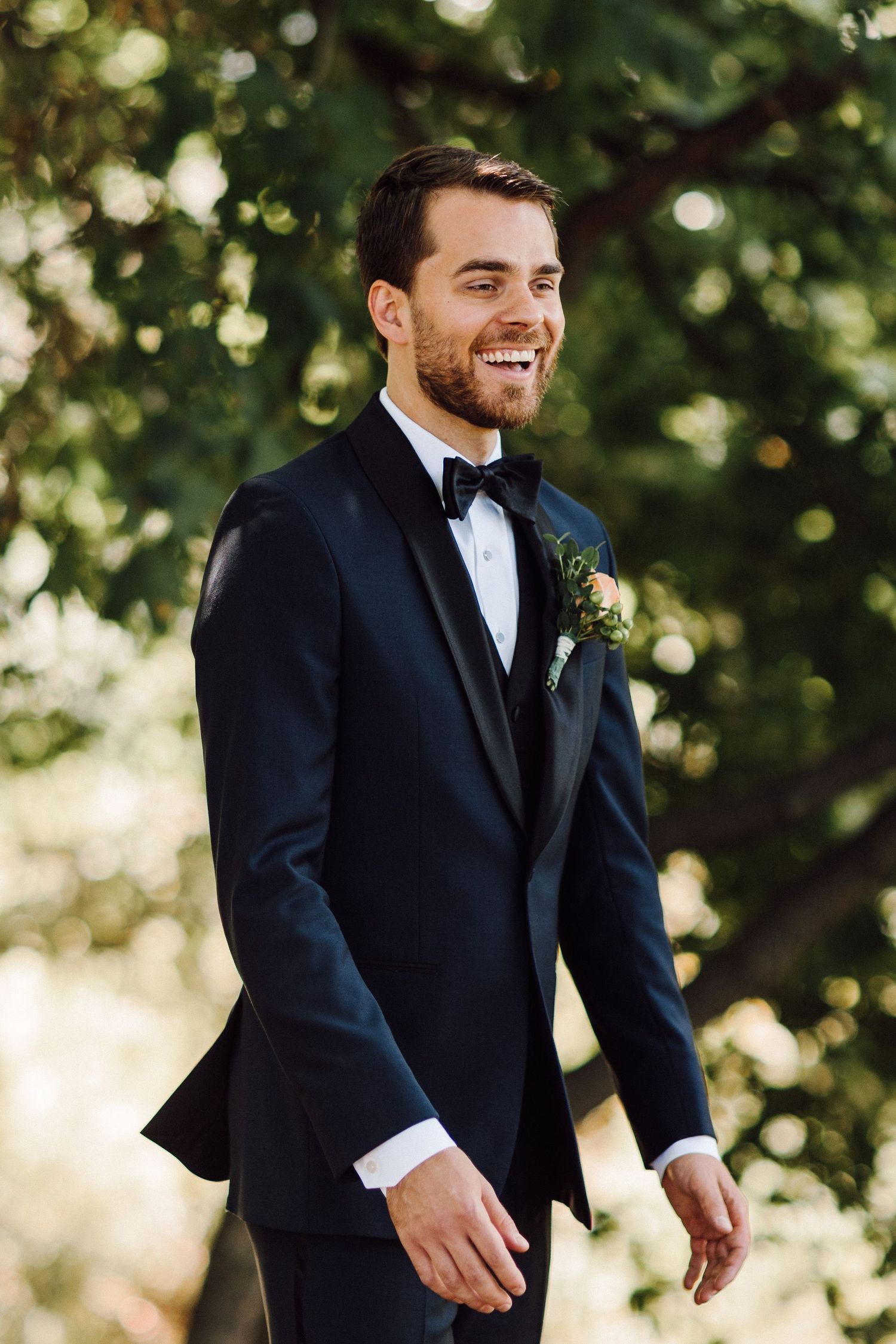 A groom in classic tuxedo smiling outdoors with natural greenery backdrop.