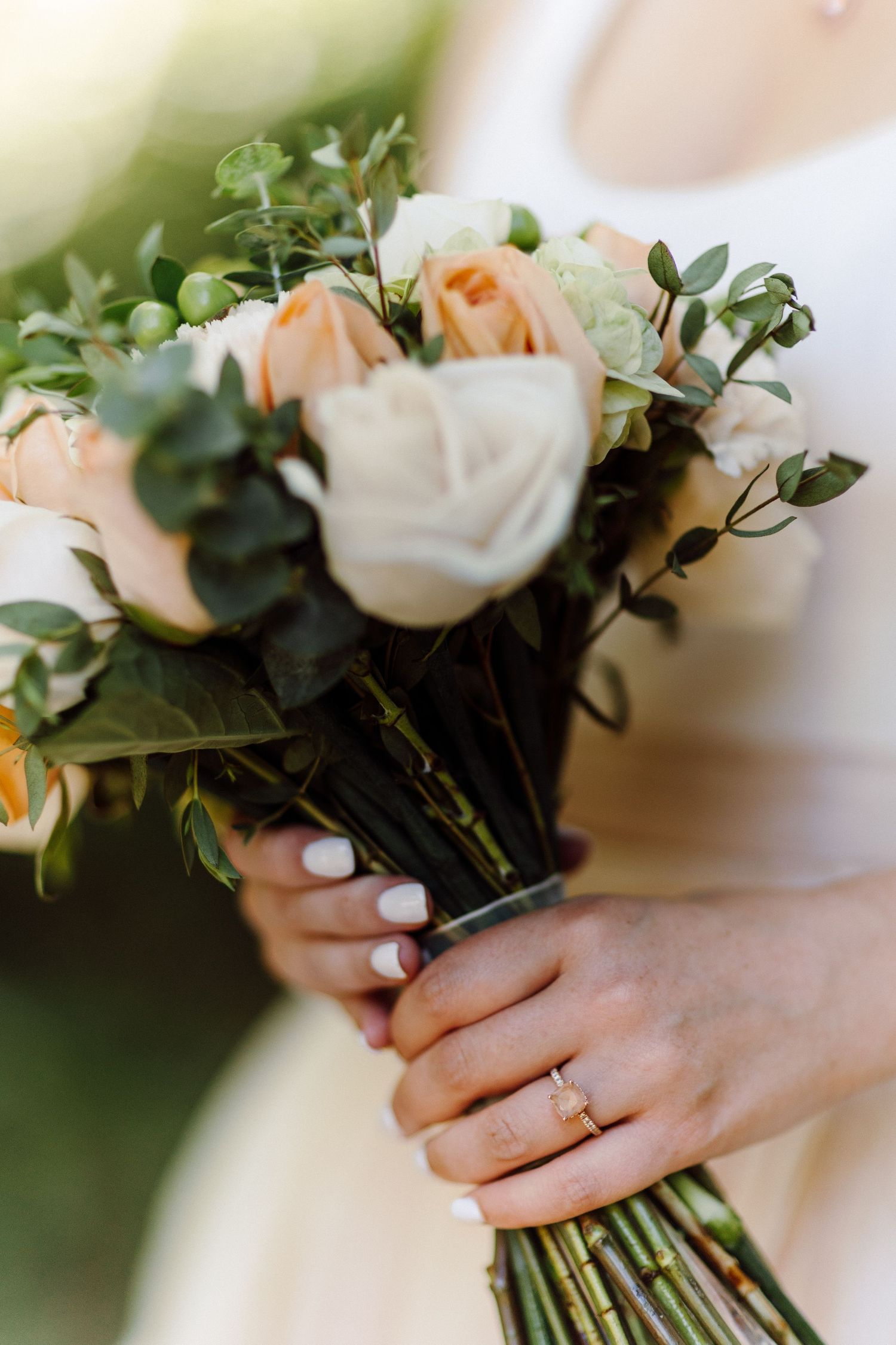 Close-up of peach and white rose wedding bouquet with delicate greenery and hands holding stems.