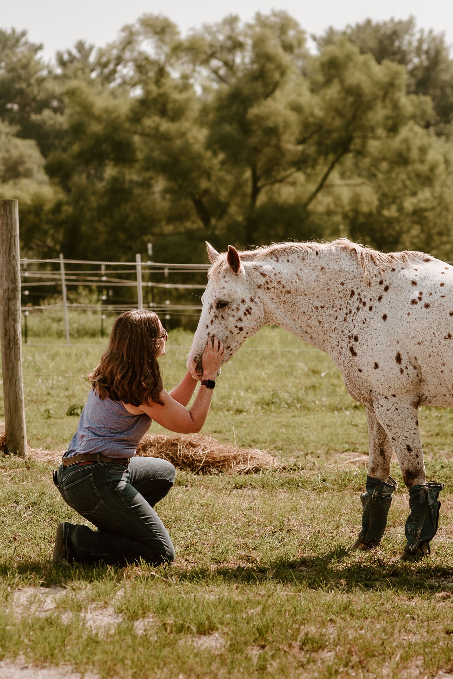 Woman in purple sleeveless shirt and denim jeans kneeling on one knee holding the muzzle of a freckled pony leaning down to her.