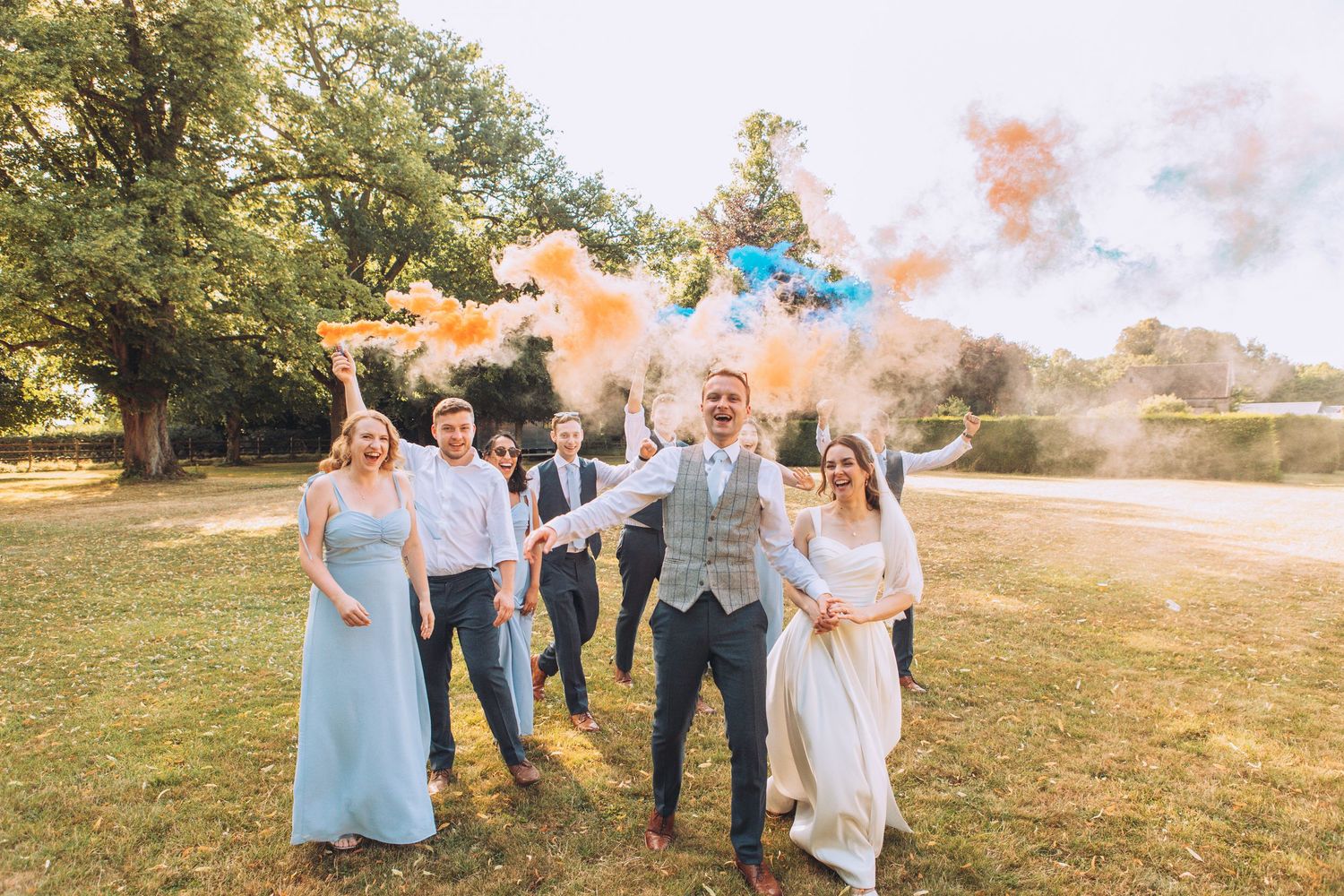 Wedding party celebrates outdoors with colorful smoke bombs in a sunny garden setting during summer festivities.