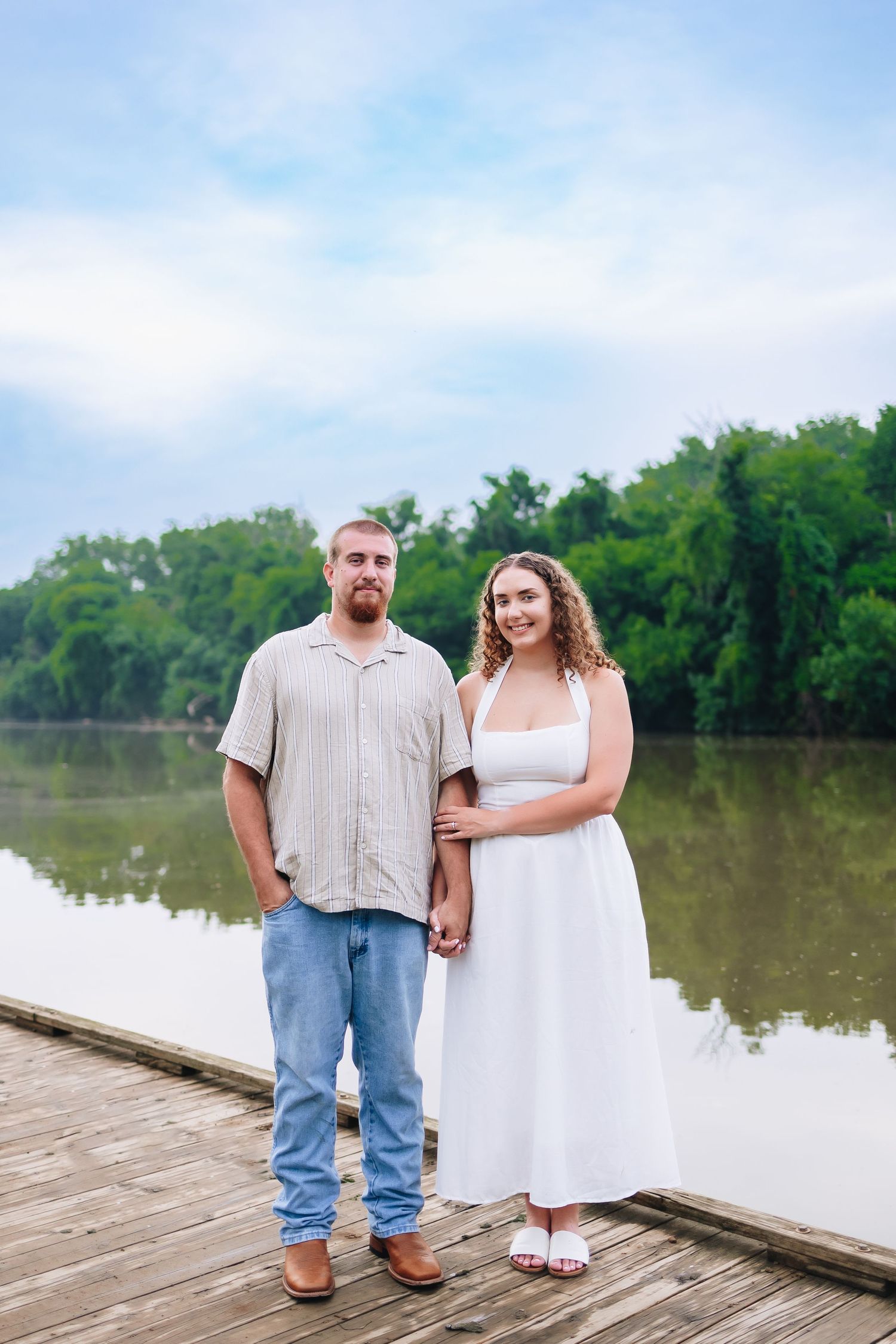 Two people stand together on a wooden dock overlooking a serene lake surrounded by trees.