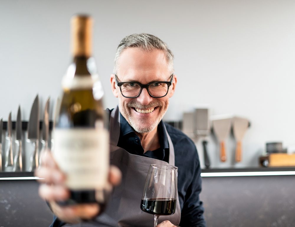 Professional in glasses holds a glass of dark wine at a counter while demonstrating wine tasting technique.