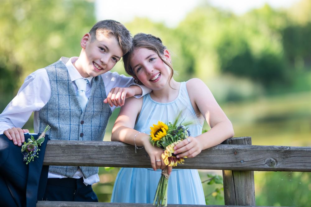 Children in formal attire lean on a wooden fence with sunflowers.