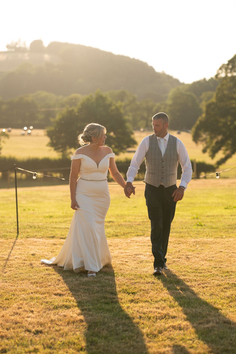 Wedding couple walking through a sun-lit field casting long shadows at sunset.