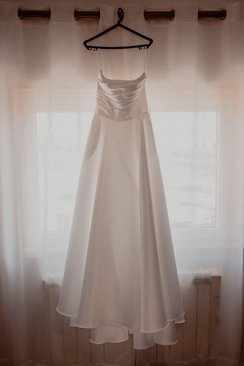 An elegant white wedding dress hangs on display against a light curtained background.
