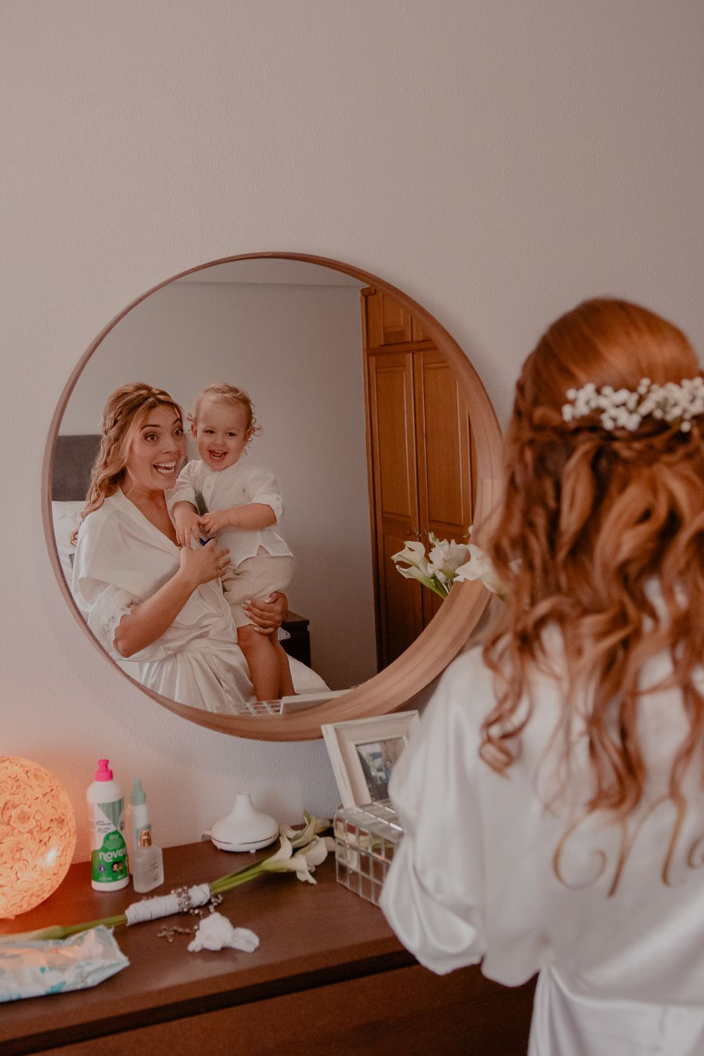 Wedding preparation scene reflected in an oval mirror with white robes and floral details.