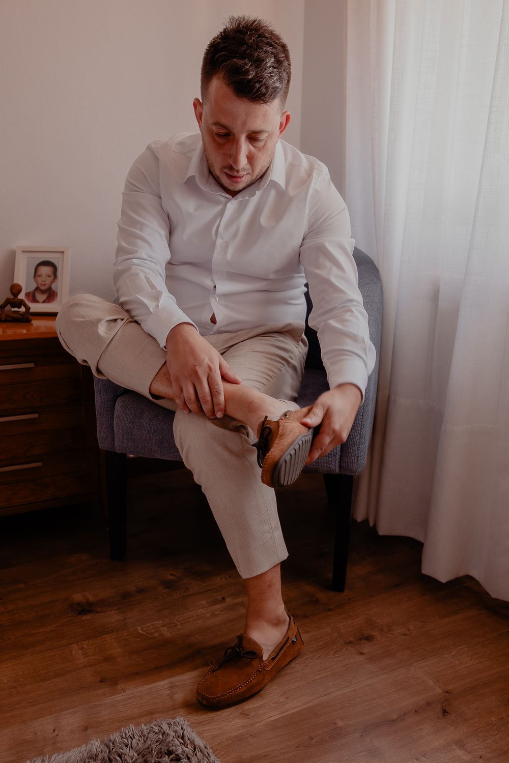 Person in white formal wear putting on dress shoes while seated on wooden floor.