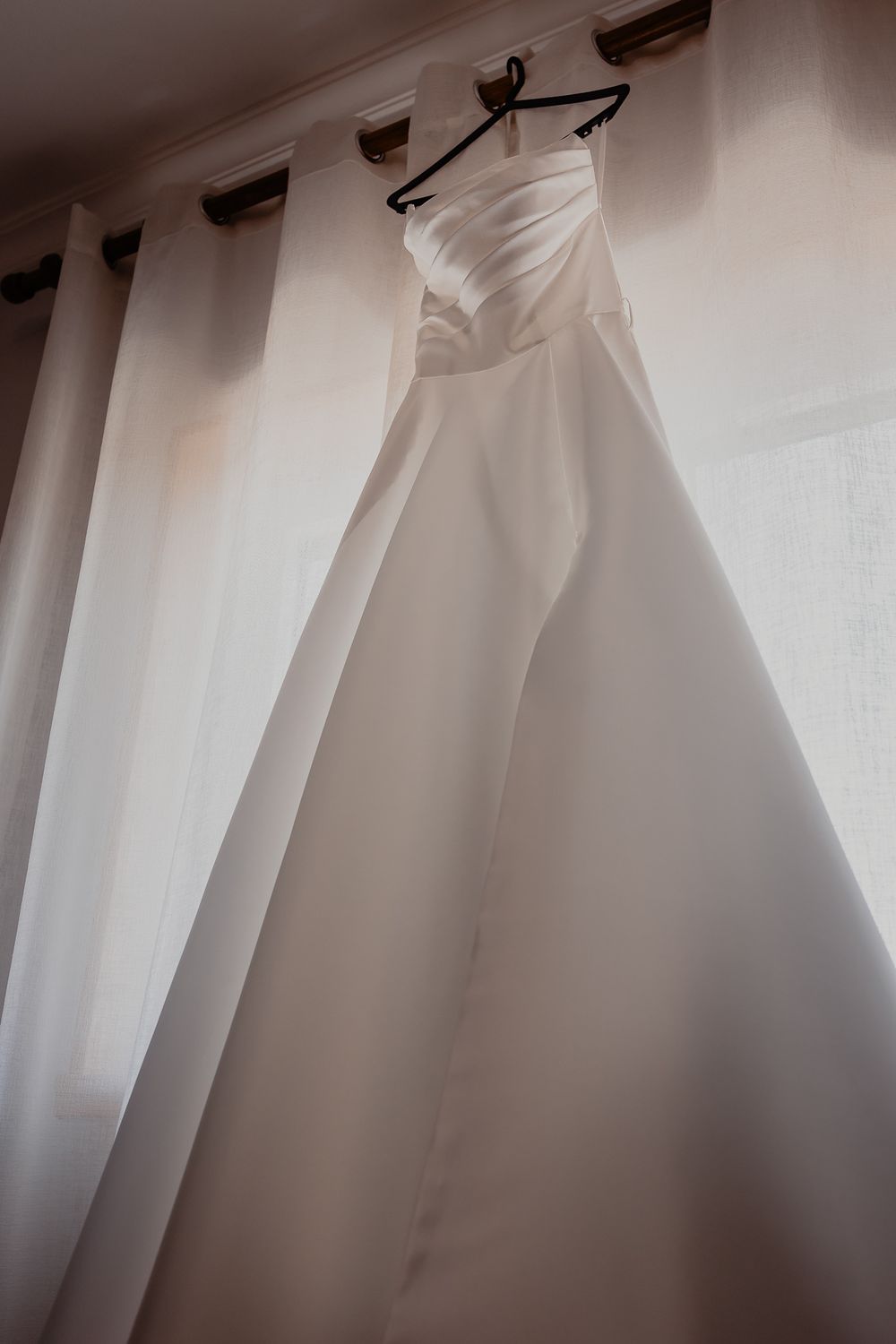A white wedding gown displayed on a hanger against soft curtains in natural light.