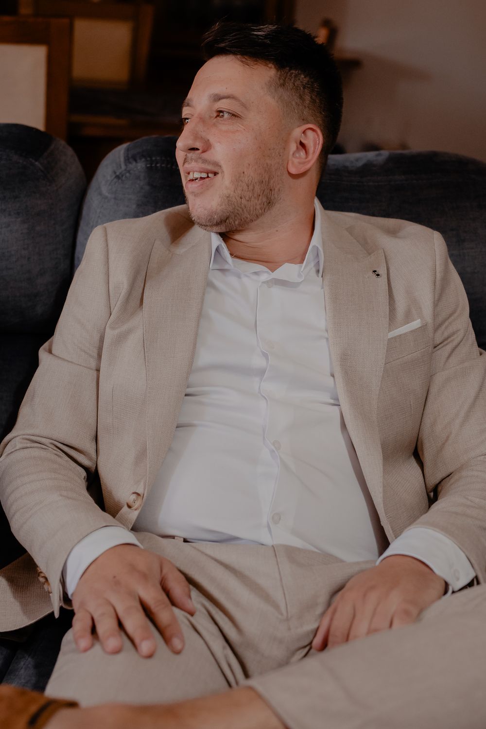 Person in beige suit and white shirt relaxing on dark couch during wedding preparations.
