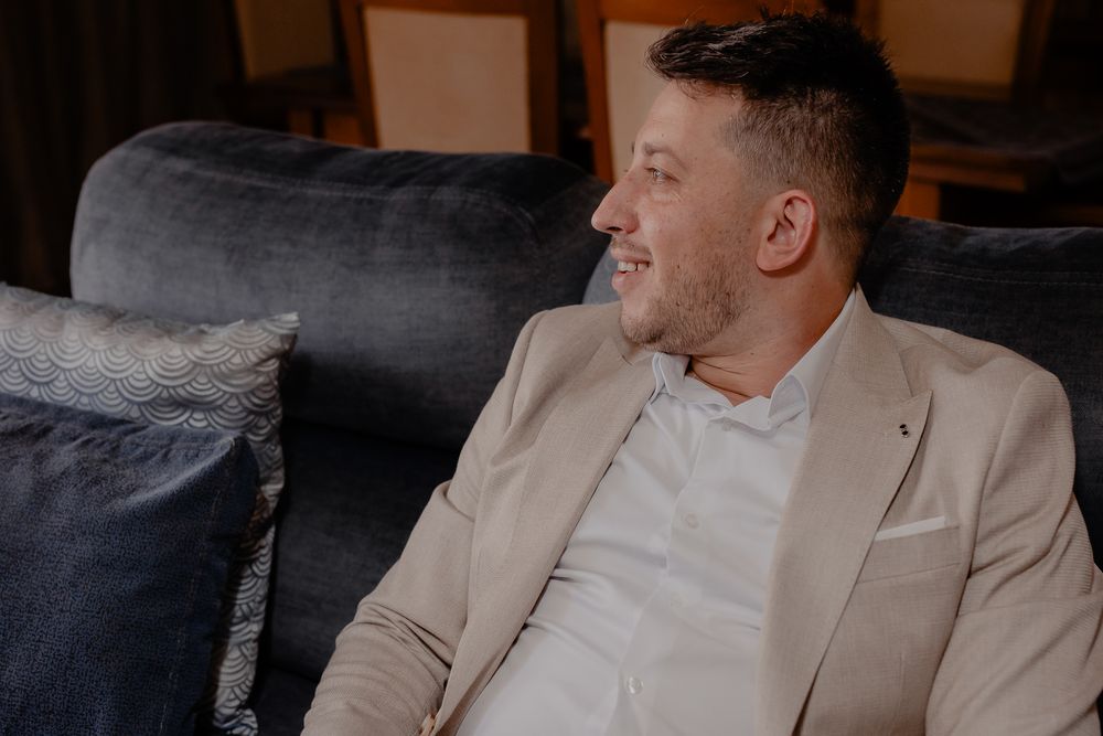 Side view of someone in beige suit sitting on dark couch during wedding event.