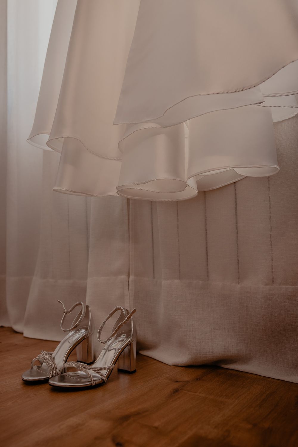 Silver bridal shoes placed beneath the flowing hem of a wedding dress on hardwood flooring.