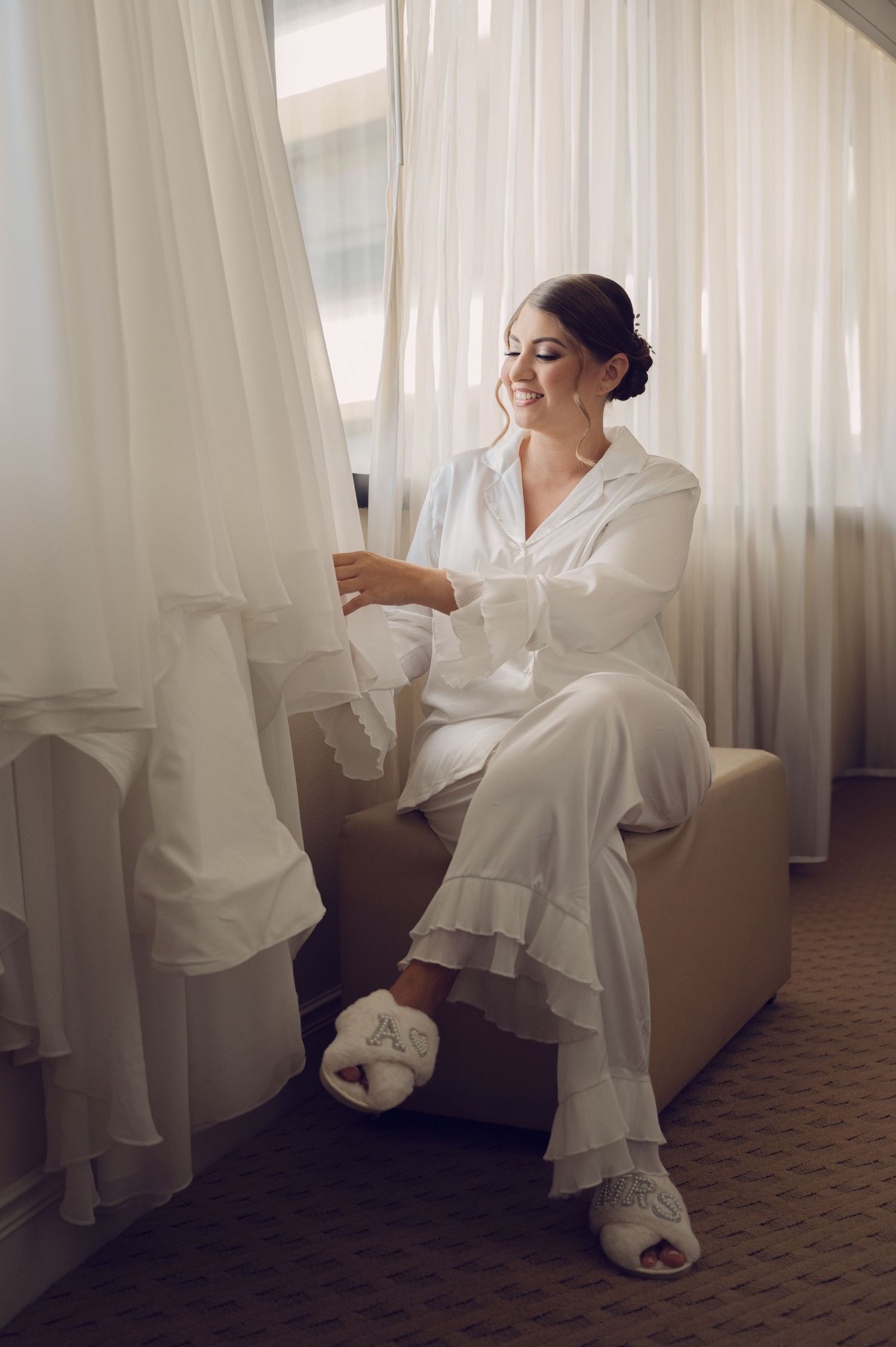 Getting ready scene with bride in white silk robe seated by window.