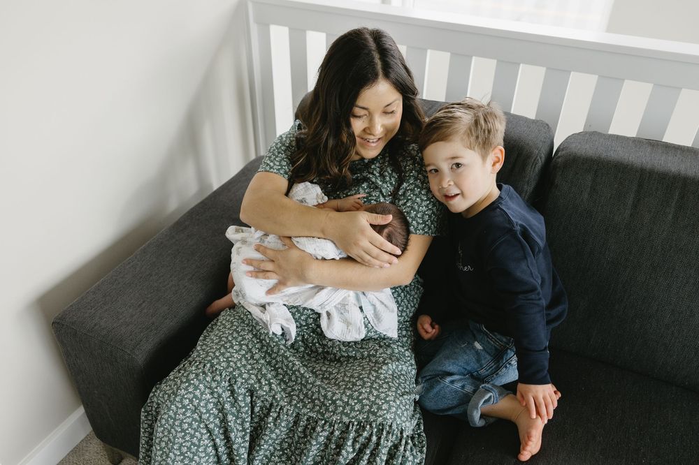 A parent sits on a grey couch with a young child and infant baby in a polka dot dress at home.