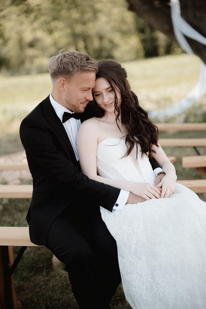 A romantic wedding couple embraces tenderly in an outdoor garden setting with wooden benches visible in the background.