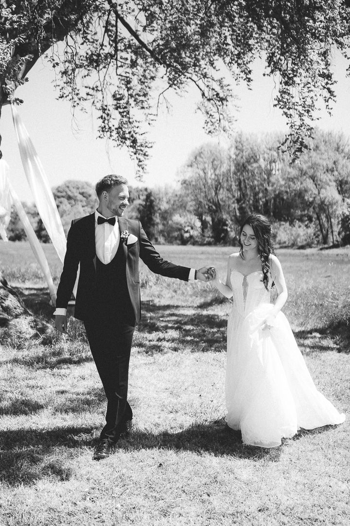 A wedding party in black and white poses playfully in a lush garden setting with trees in the background.