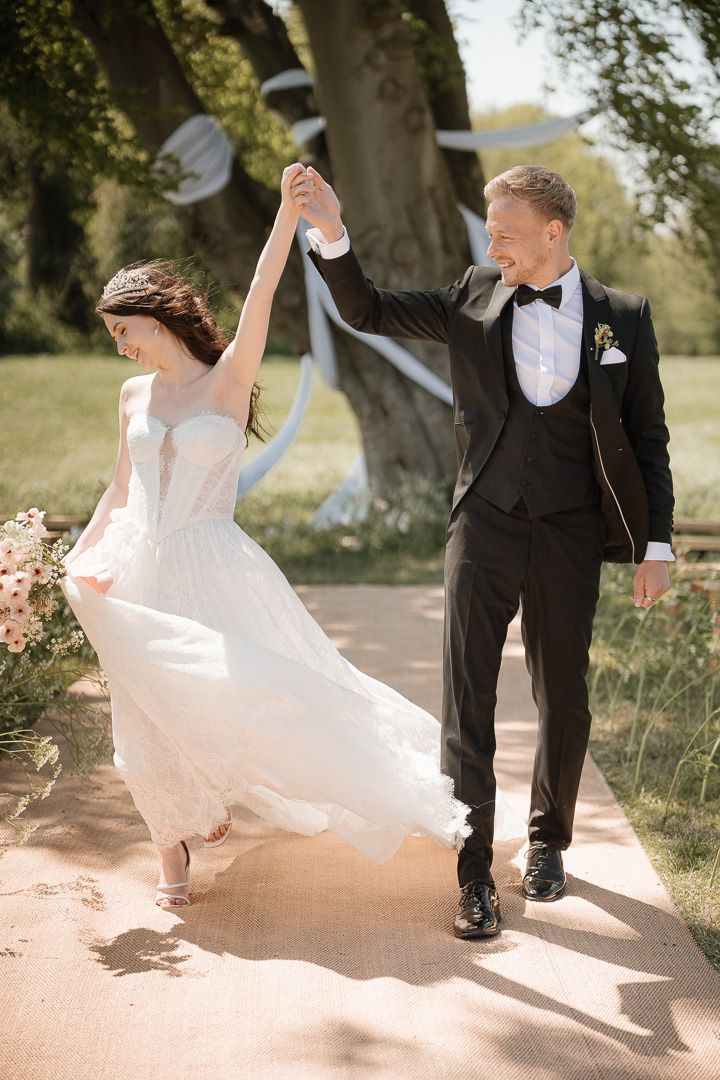 A wedding couple dances joyfully on a garden path as sunlight filters through trees in the background.