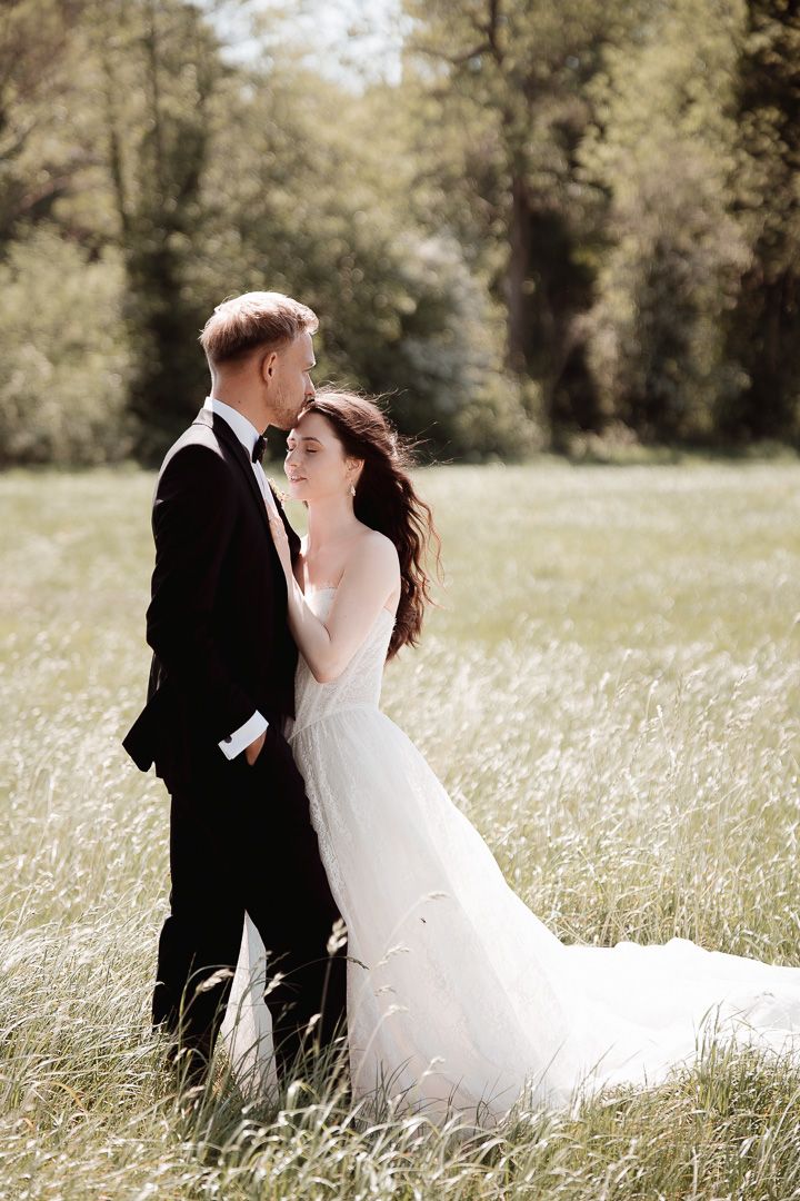 A romantic couple in wedding attire embraces in a sunlit meadow filled with tall grass and flowers.