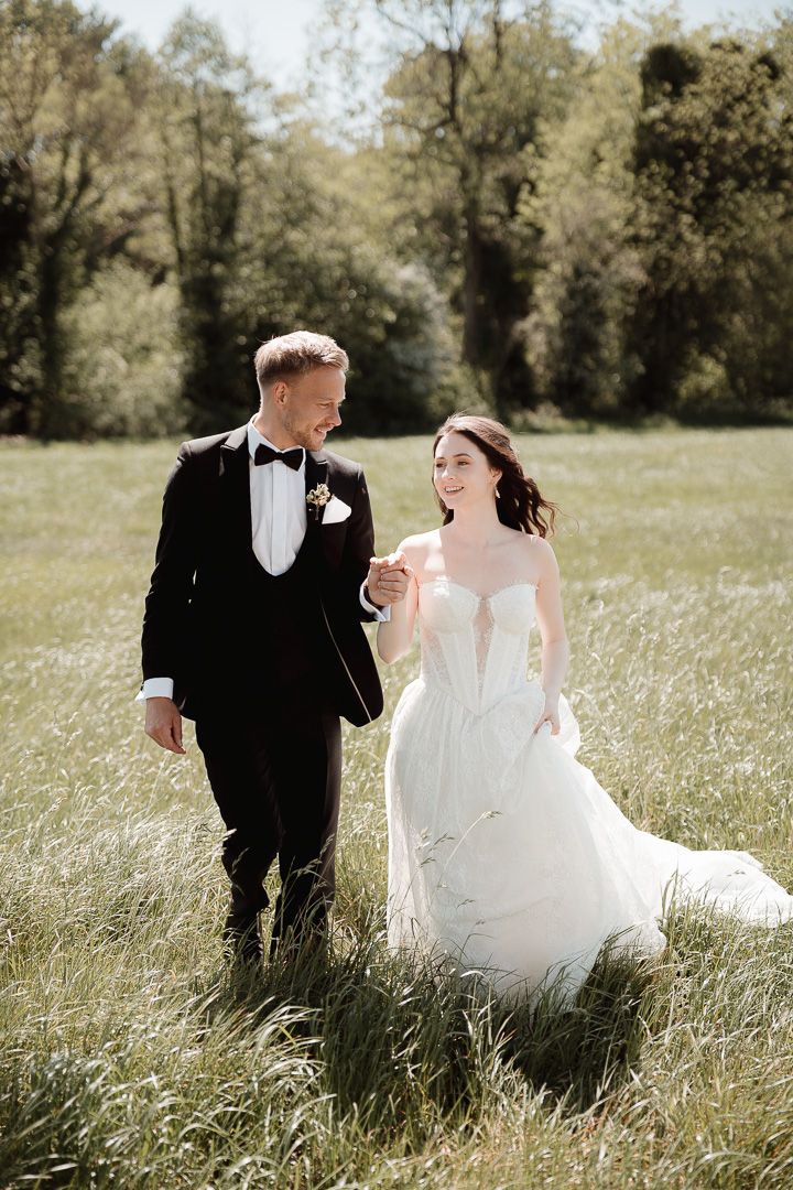 A newlywed couple strolls through a sunny meadow, with the bride in white and groom in black tuxedo.