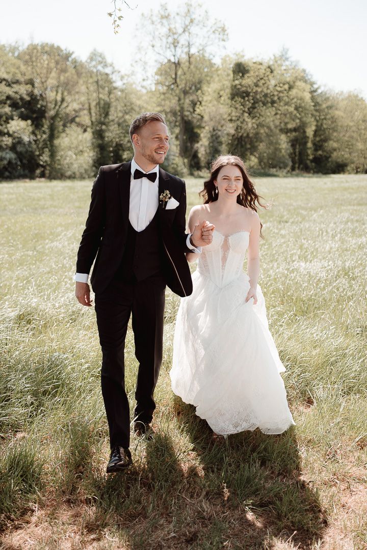 Newlywed couple walking together through a sunlit meadow with trees in background.