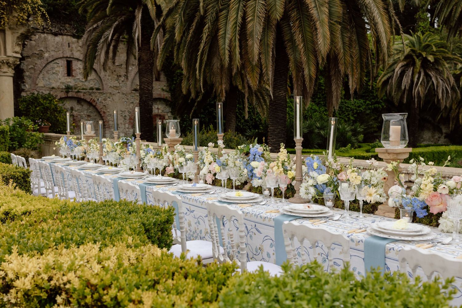 An elegant outdoor wedding reception table set with white linens and floral centerpieces under palm trees in a garden setting.