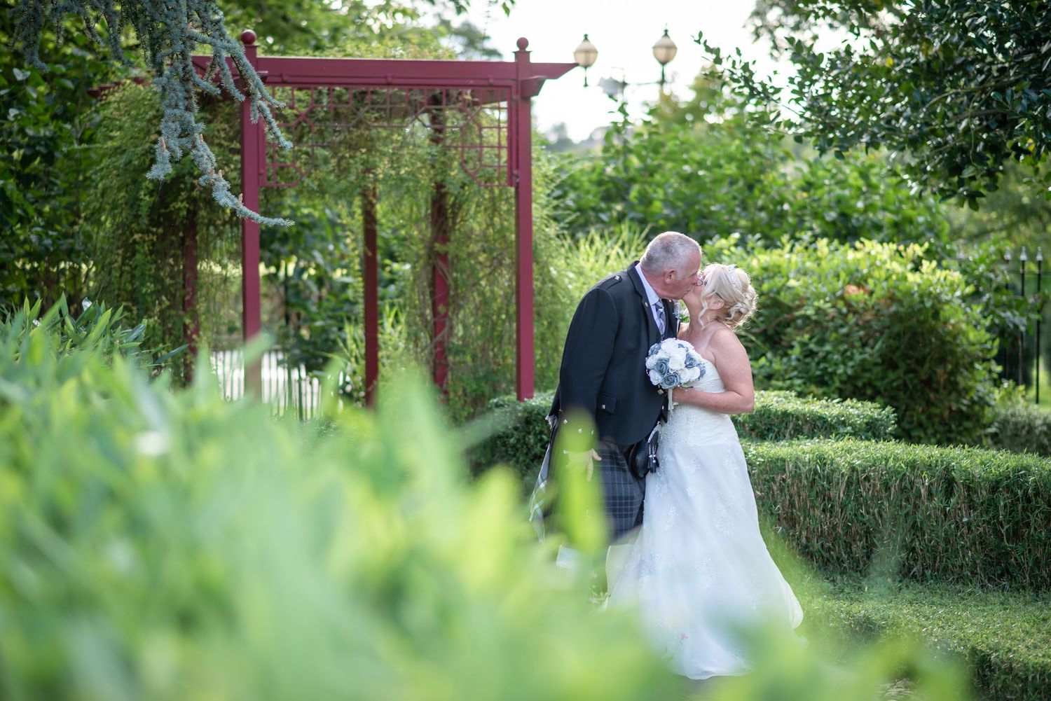 A romantic scene in a lush garden with a red arbor pergola surrounded by green hedges and foliage.
