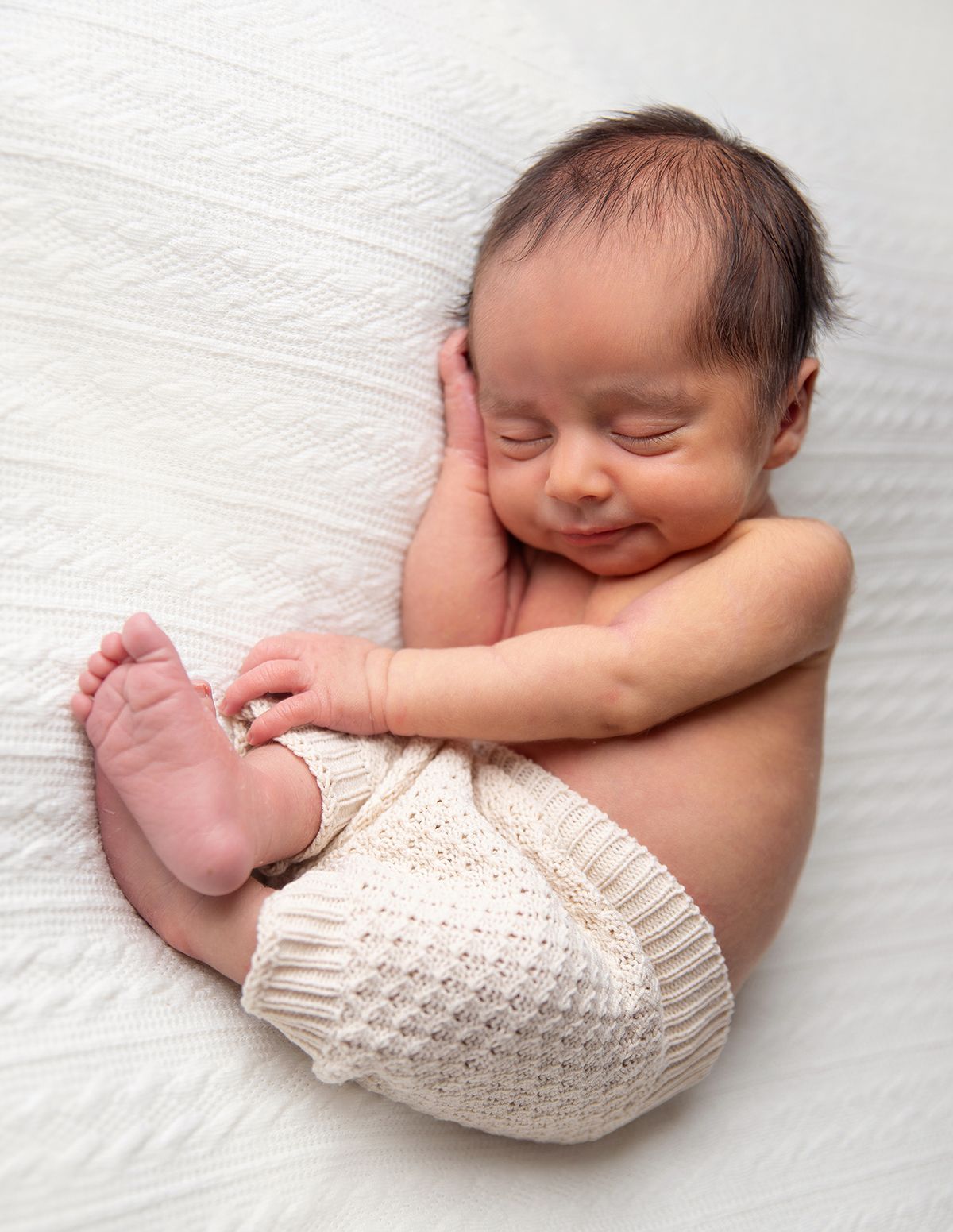 Sleeping newborn with knitted pants curls up peacefully against soft white textured bedding. During a newborn session in Bexley