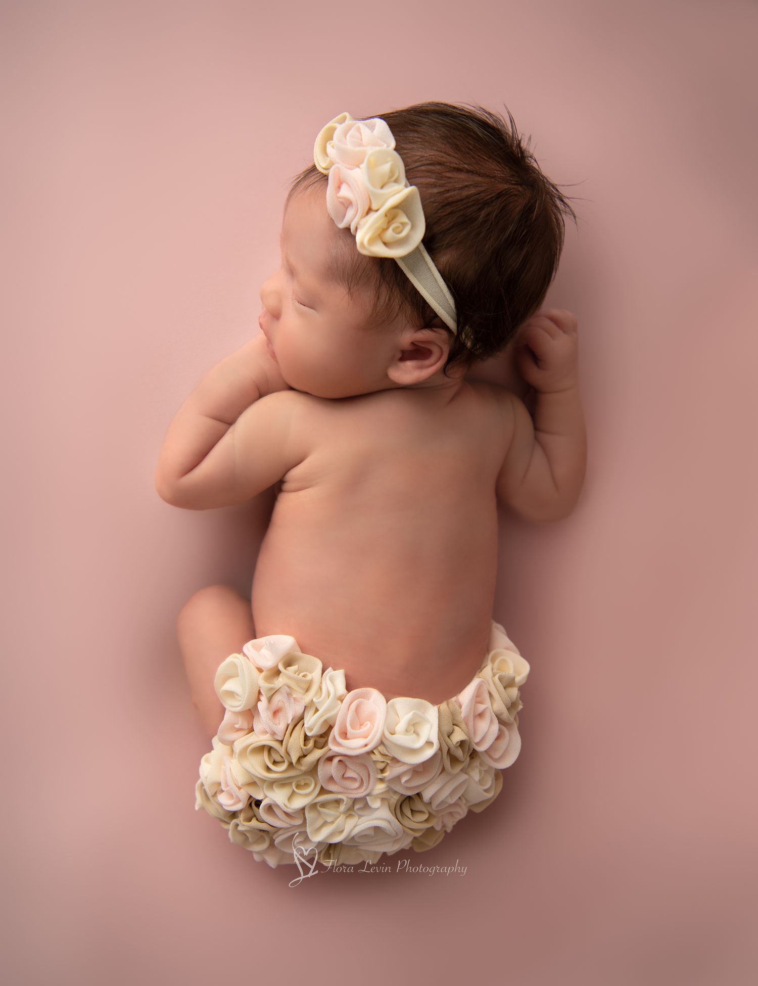 Newborn baby sleeping on pink backdrop with white and neutral floral pants on her tummy