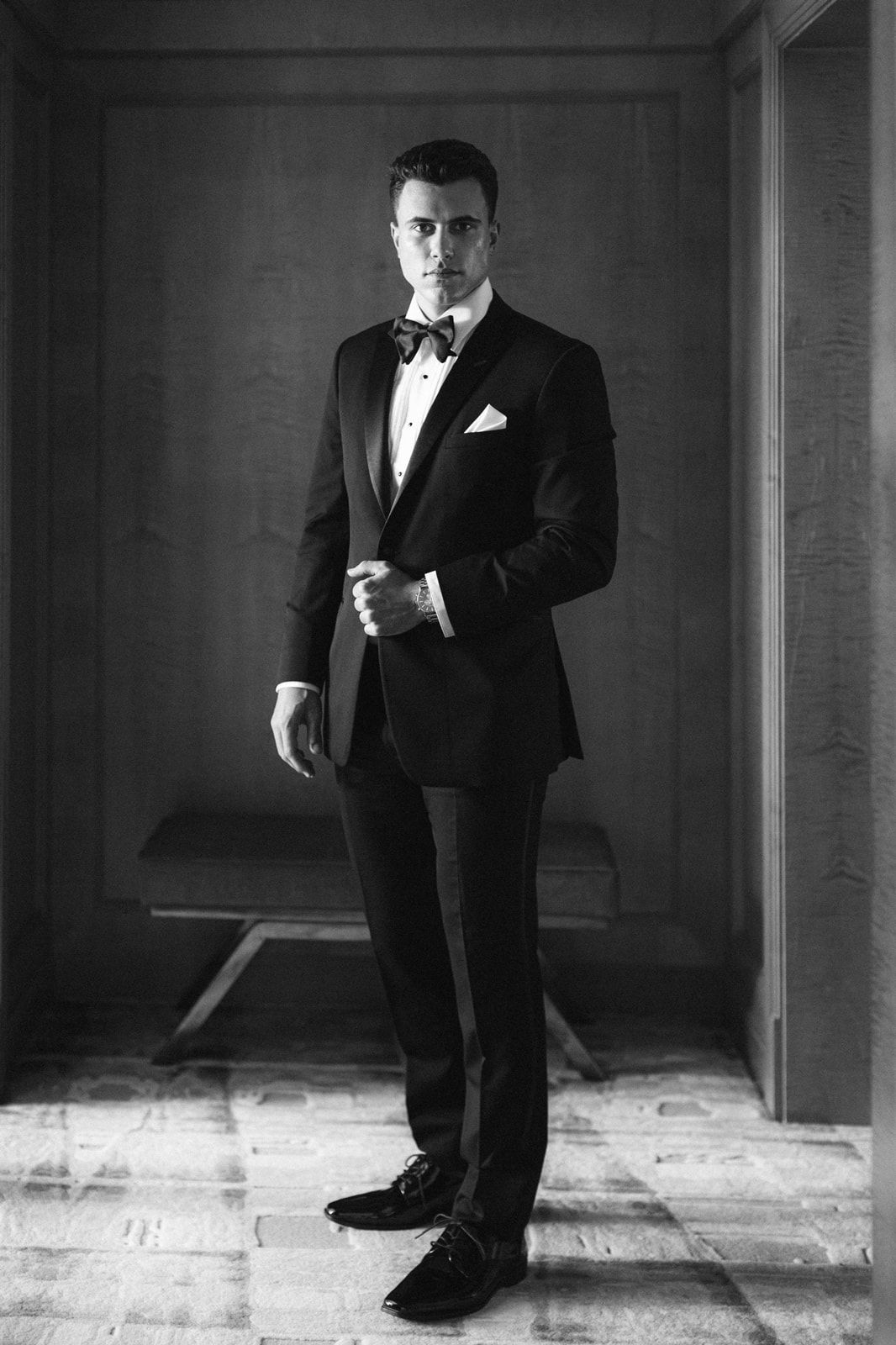 A black and white portrait of someone in a classic tuxedo with bow tie standing in an elegant indoor setting.